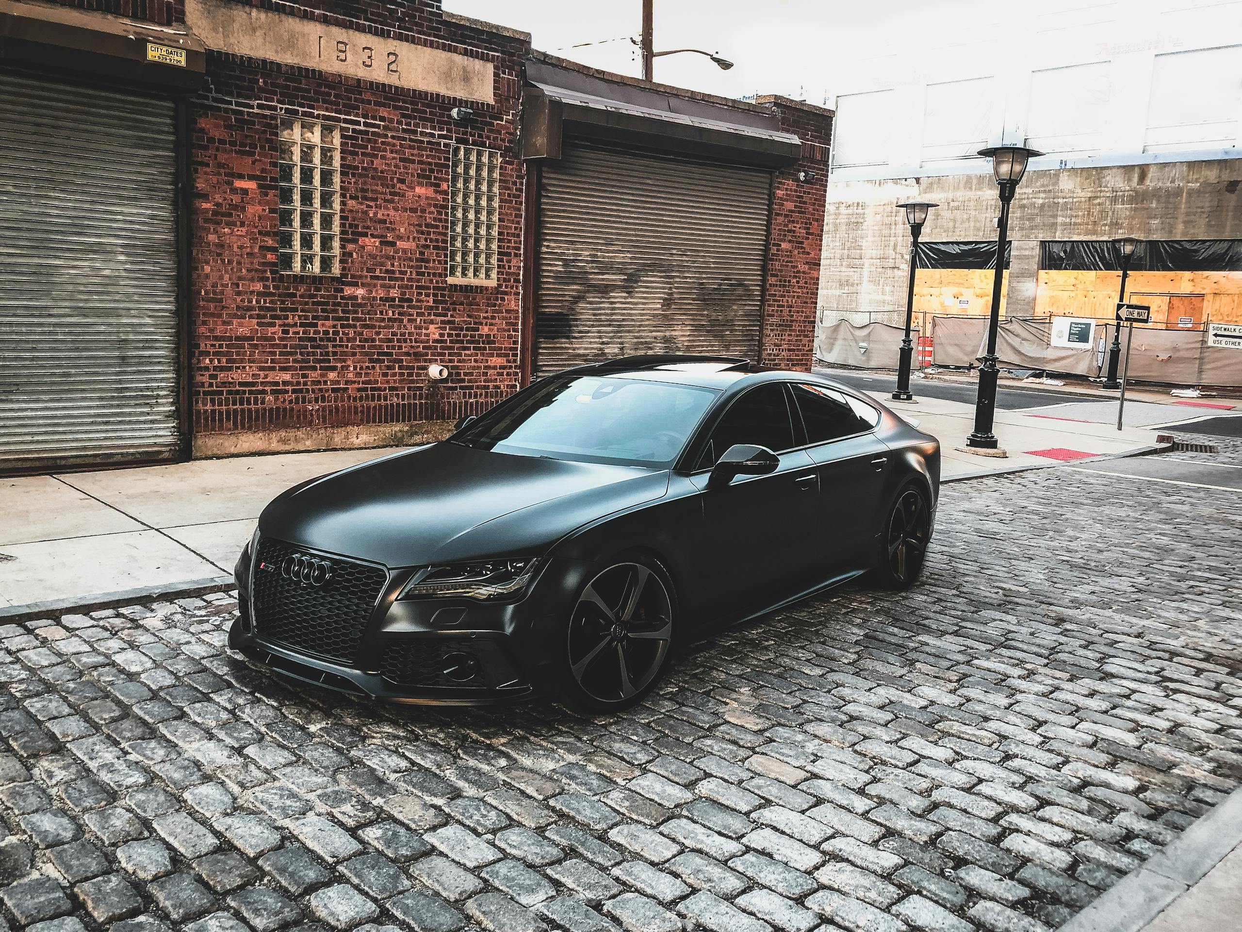 A sleek black car parked on a cobblestone street in an urban setting, featuring brick buildings.