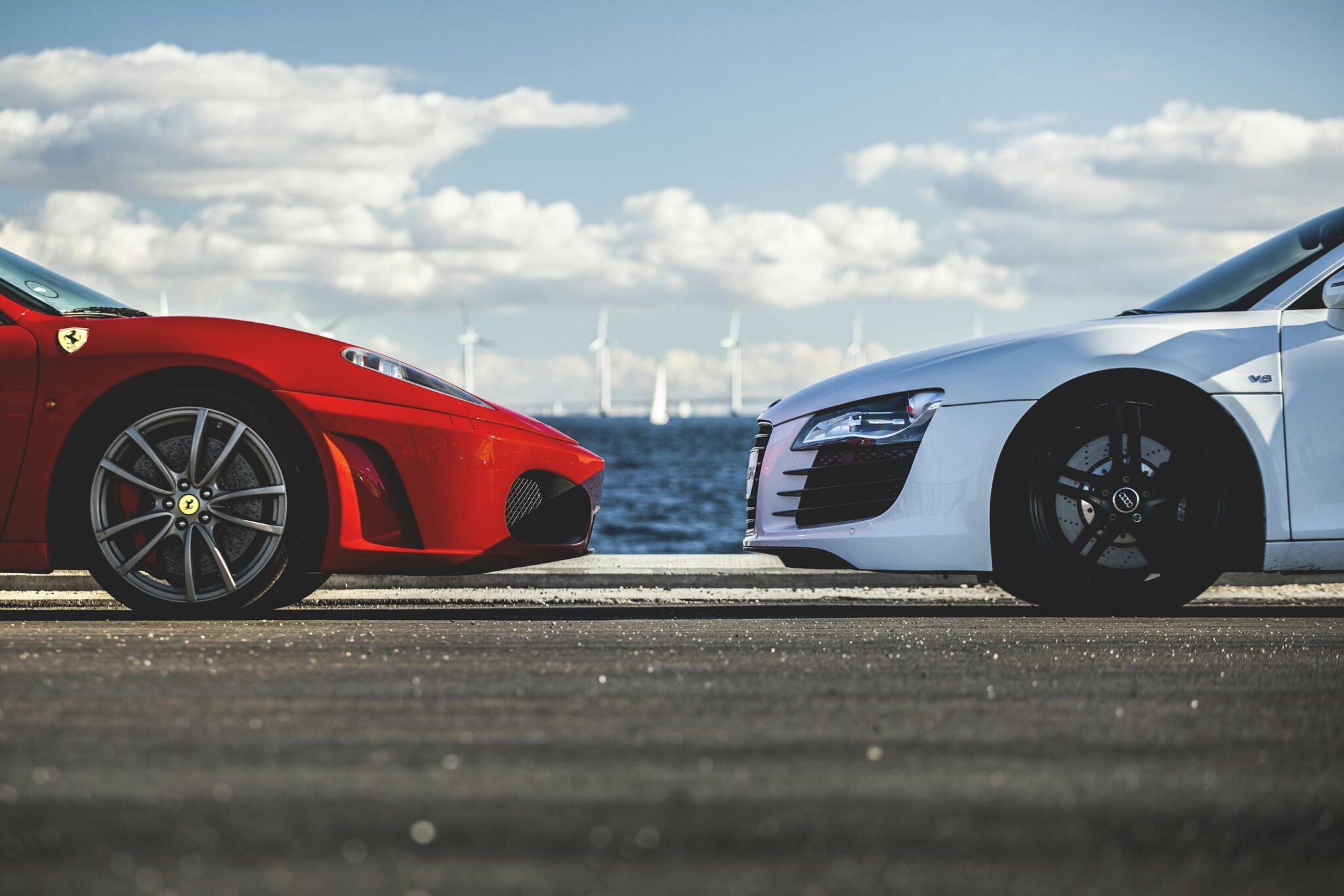 Red Ferrari and white Audi R8 facing each other with wind turbines in the background by the sea.